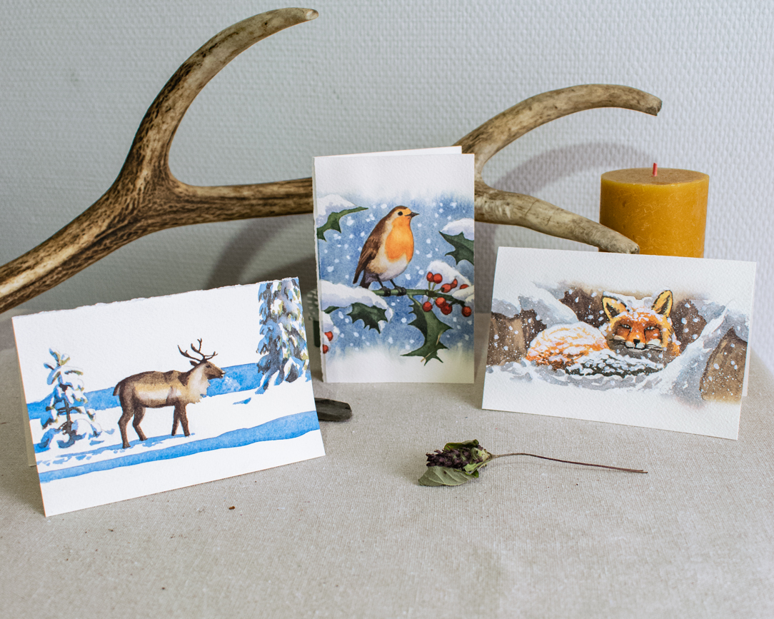 Trois cartes de vœux sont posées côte à côte sur une toile de lin naturel. Une fleur séchée est posée sur le devant de la présentation, tandis qu'au fond, il y a un bois de cerf, une bougie orange et une petite pierre grise. Le mur derrière est blanc et texturé.