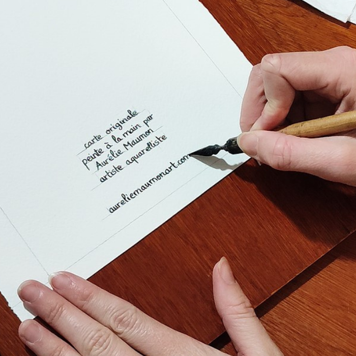 Une main tenant une plume à écriture est en train de tracer les les lettres au dos d'une carte de vœux. Le texte est tracé à l'encre de Chine et dit : "carte originale peinte à la main par Aurélie Maumon artiste aquarelliste aureliemaumonart.com"