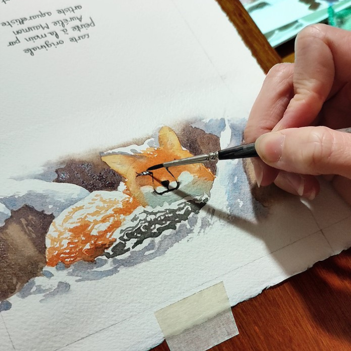 Une main tenant un petit pinceau est en train de tracer les yeux d'un renard à l'aquarelle. La peinture est une aquarelle sur un papier en coton représentant un renard couché sous la neige qui tombe à gros flocons.