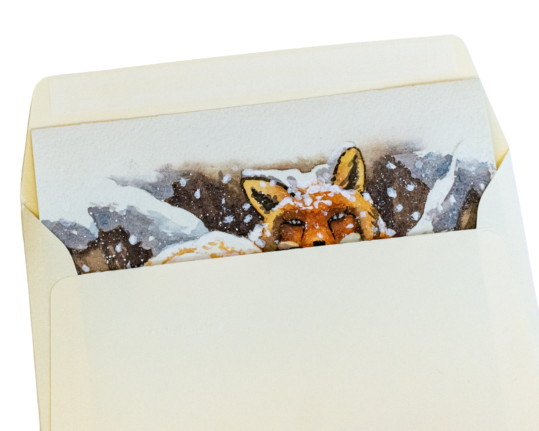 Une carte de vœux est glissée dans une enveloppe. La carte de vœux peinte à la main représente un renard couché dans la neige avec de gros flocons qui tombent. L'enveloppe est de couleur ivoire.