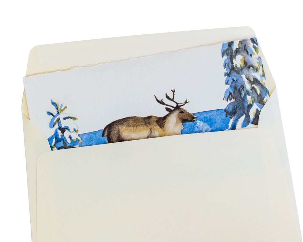 Une carte de vœux est glissée dans une enveloppe. La carte est peinte à la main et représente un renne marchant dans la neige au soleil et à côté d'épicéas couverts d'une épaisse couche de neige. L'ombre des arbres s'étend dans une couleur bleue vive et on voit la brume causée par le souffle du renne. L'enveloppe est de couleur ivoire.