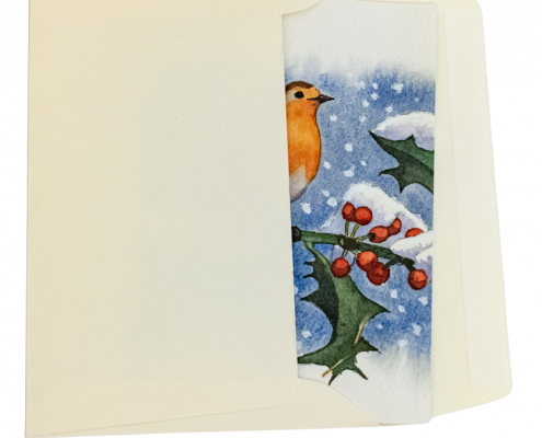 Une carte de vœux est glissée dans une enveloppe. La carte de vœux est peinte à la main et représente un rouge-gorge posé sur une branche de houx. Les feuilles et les baies du houx sont couvertes de neige. Le fond de la carte est bleue outremer avec des flocons de neige qui tombent. L'enveloppe est de couleur ivoire.